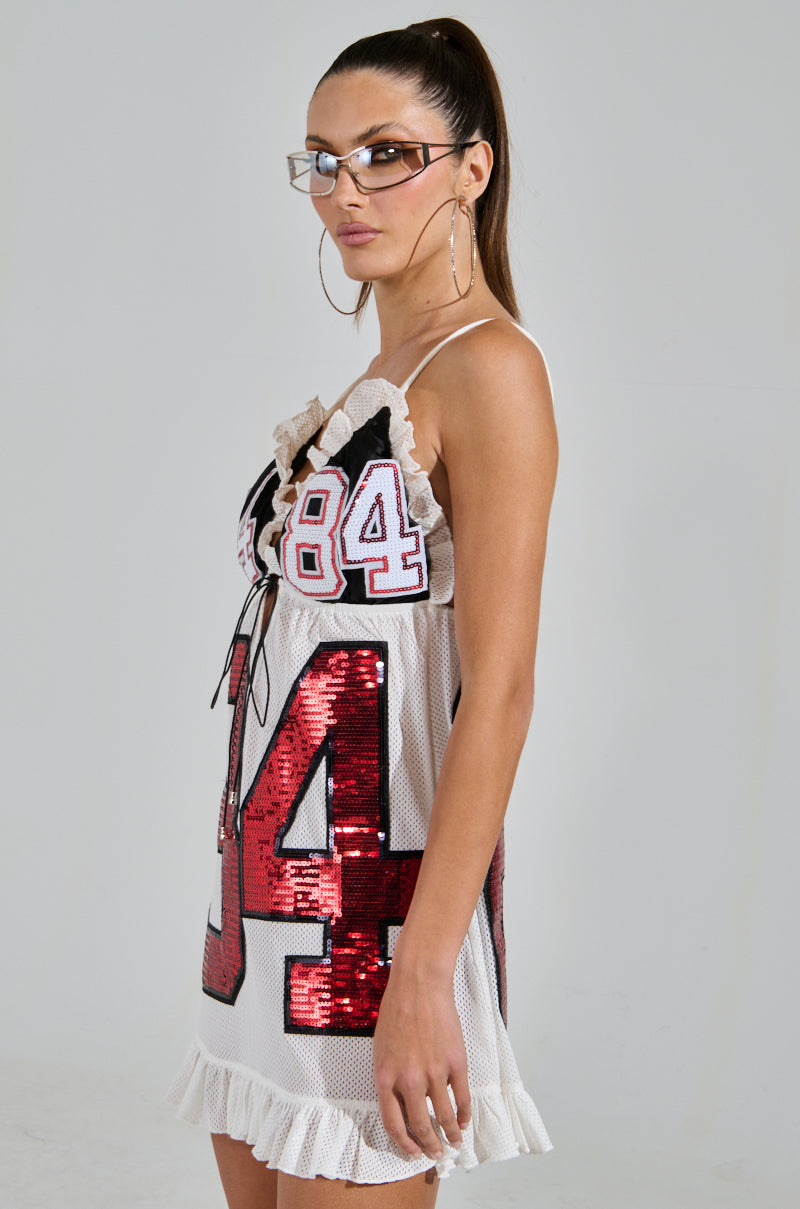 A woman with long hair in a ponytail wears glasses, hoop earrings, and a sleeveless TOUCHDOWN SEQUIN JERSEY DRESS featuring large "84" sequin patches. The football-inspired dress has ruffled details at the top and hem as she stands against a plain background.