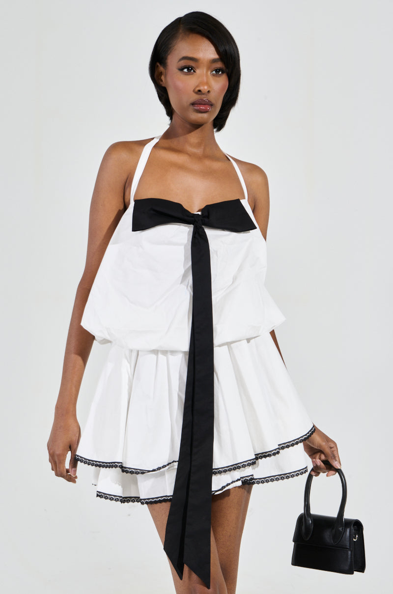 A woman in a white MIA POPLIN MINI DRESS WITH BOW, adorned with a black bow applique and trim, holds a small black handbag. She stands against a plain white background, looking forward.