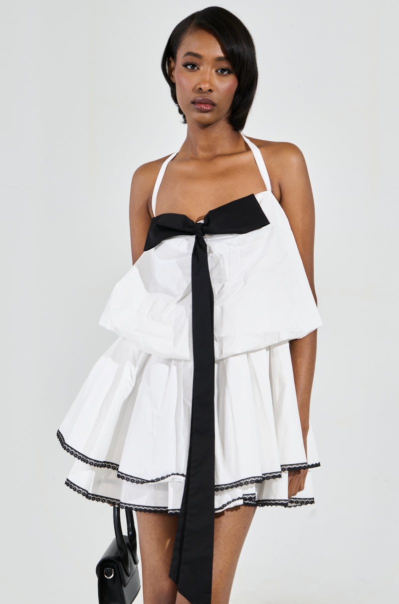 A woman is dressed in the MIA POPLIN MINI DRESS WITH BOW, a white skater dress adorned with a large black bow applique on the front. It features multiple layers and black trim. Her straight dark hair contrasts against a plain backdrop.