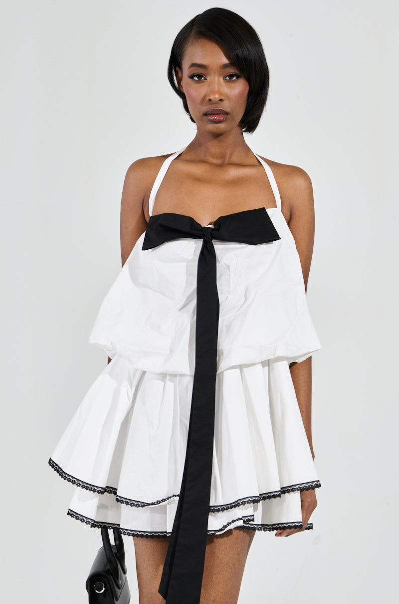 A person poses against a plain background wearing the MIA POPLIN MINI DRESS WITH BOW, featuring a white strapless design adorned with a large black bow applique on the front and accented with black trim along the hem. They hold a sleek black handbag at their side.
