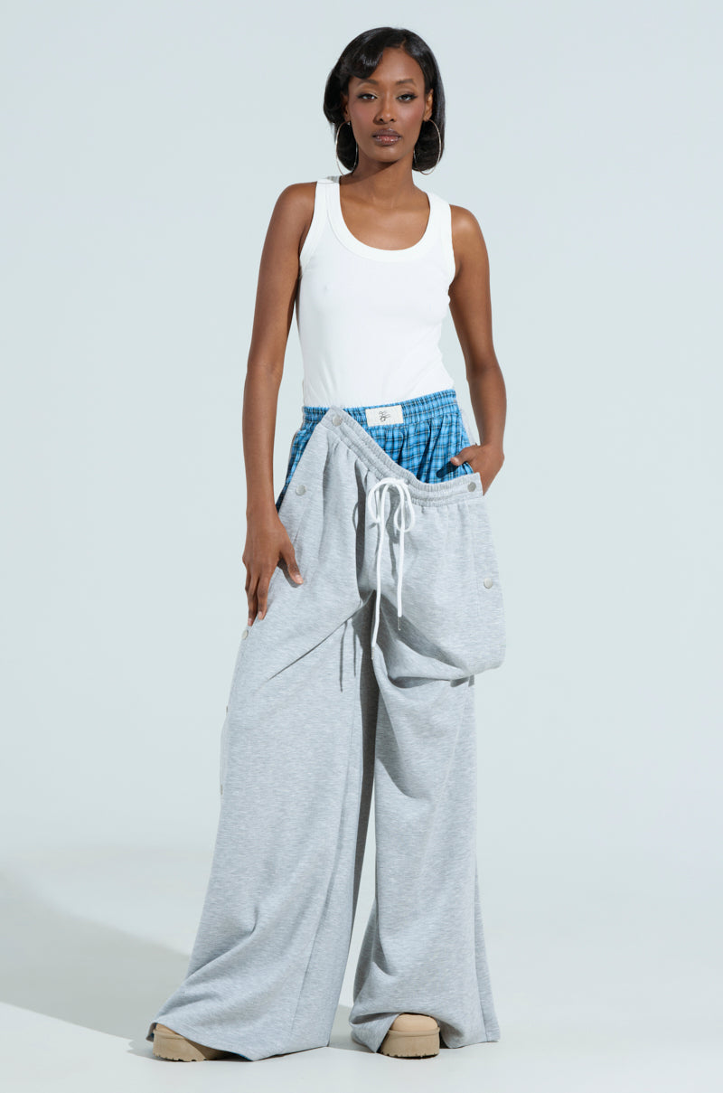 A person stands confidently against a light gray background, wearing the RUNNING LATE JUMPSUIT with a mixed pattern design and beige platform shoes. Their right hand is in their pocket, and they have a focused expression.
