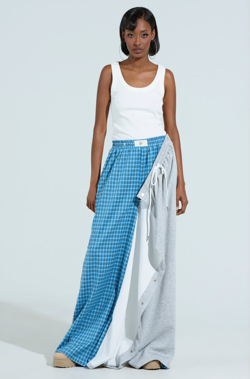 A woman stands against a plain background wearing the RUNNING LATE JUMPSUIT, a white tank top with unique trousers featuring a pattern design of blue plaid and gray fabric, exuding an effortless style.