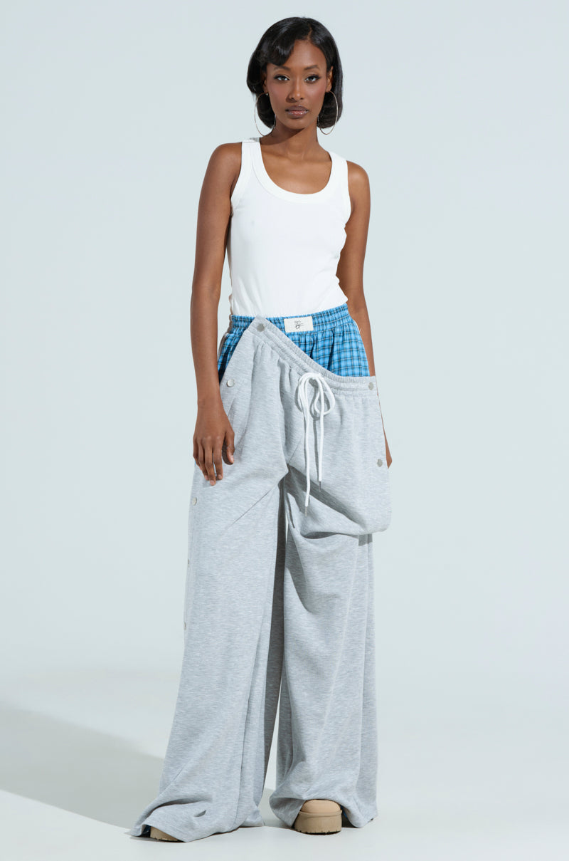 Dressed in a stylish mix, someone confidently poses in the RUNNING LATE JUMPSUIT, featuring a white tank-style top with gray wide-leg pants and blue plaid boxers peeking out. With short, styled hair, they embrace the look against a plain backdrop.