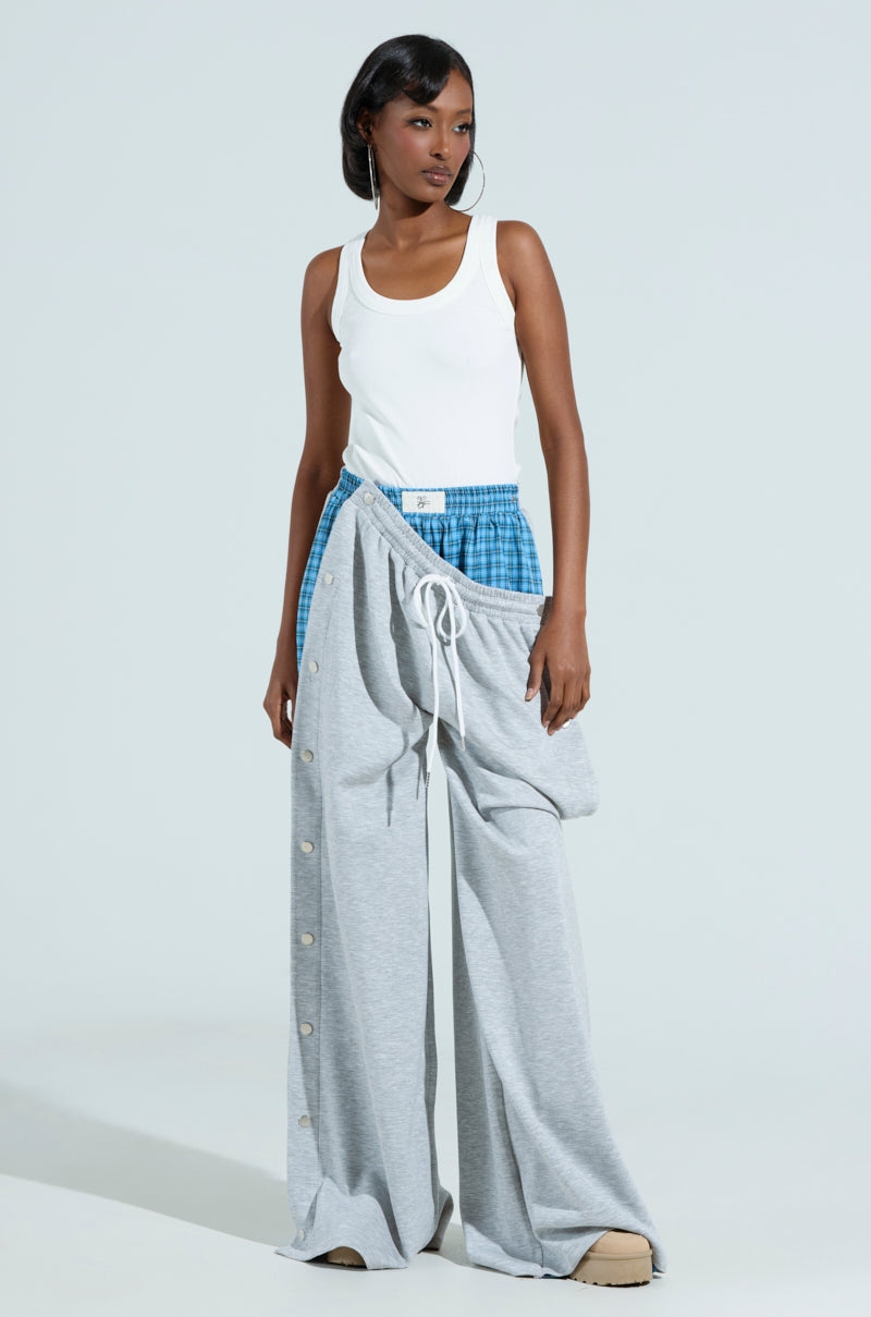 Against a plain backdrop, a person exudes confidence in the RUNNING LATE JUMPSUIT with its ribbed knit design, creatively layered over oversized gray pants featuring button details. Their short dark hair perfectly complements the mixed pattern style.