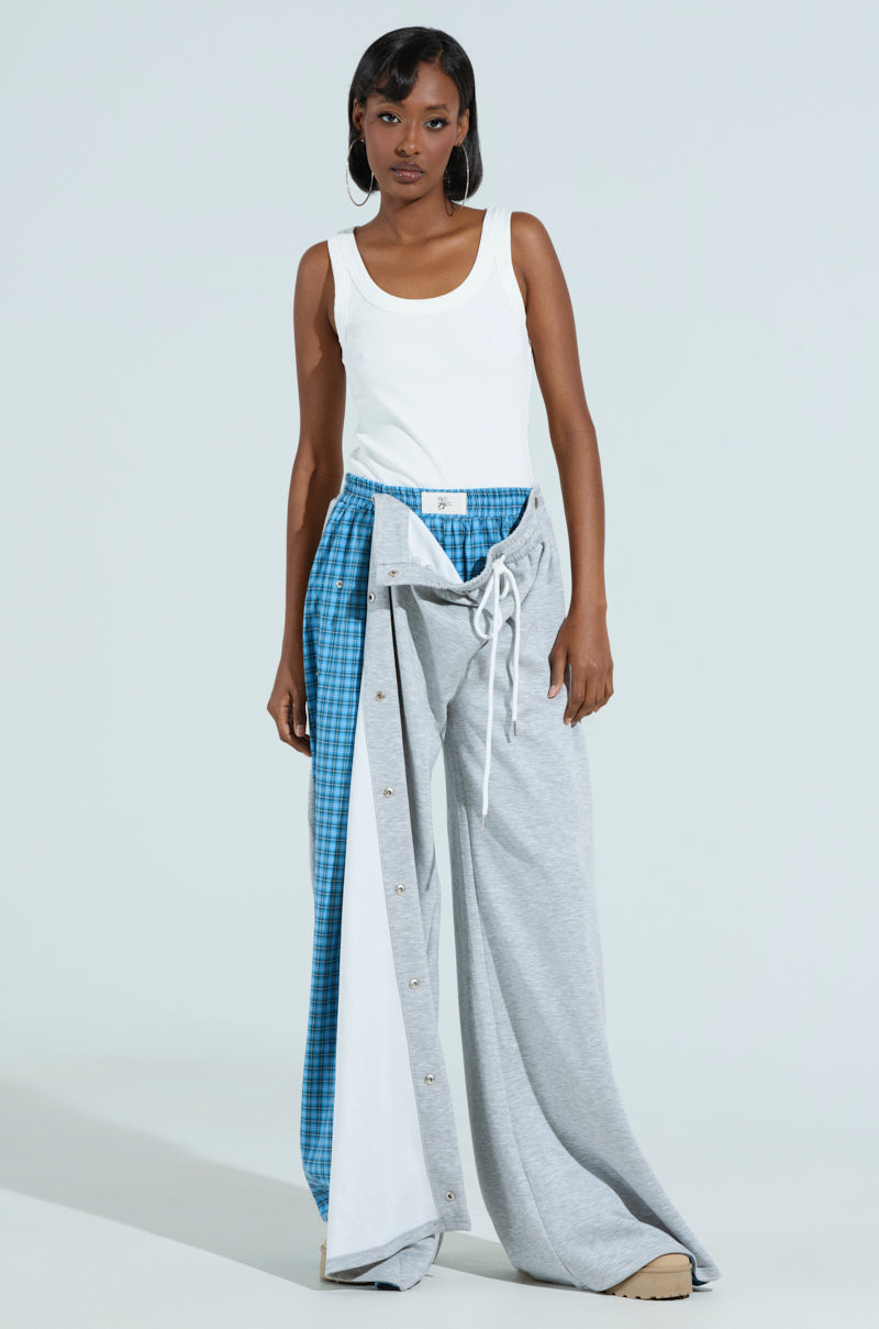 A woman stands against a plain background, wearing the RUNNING LATE JUMPSUIT, blending plaid wide-leg style with grey sweatpants. She accessorizes with hoop earrings and tan sneakers, her hand casually resting in a pocket.