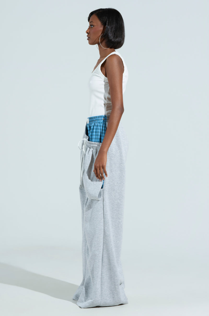 A person stands in profile against a plain background, showcasing the RUNNING LATE JUMPSUIT with its ribbed knit top and stylish wide plaid pants featuring chic blue waist detailing.