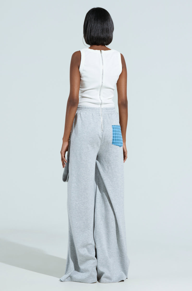 A person with black hair is seen from behind wearing the RUNNING LATE JUMPSUIT, a white sleeveless top with wide-leg gray pants and a blue checkered pocket patch. The plain light gray background highlights the jumpsuit's mixed pattern design seamlessly.