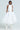 Against a plain background, a person with shoulder-length hair and hoop earrings wears a sleeveless white halter top with ruched detailing and THE BEST MAXI BUBBLE SKIRT in midi length. The look is completed with high-rise white footwear.