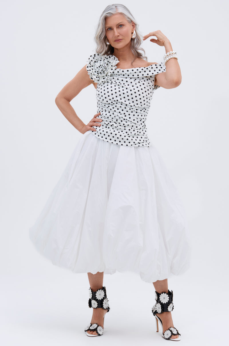 A woman with long gray hair wears a white off-shoulder polka-dot top, the THE BEST MAXI BUBBLE SKIRT, and high-heeled sandals with large white flowers, posing confidently against a plain white background.