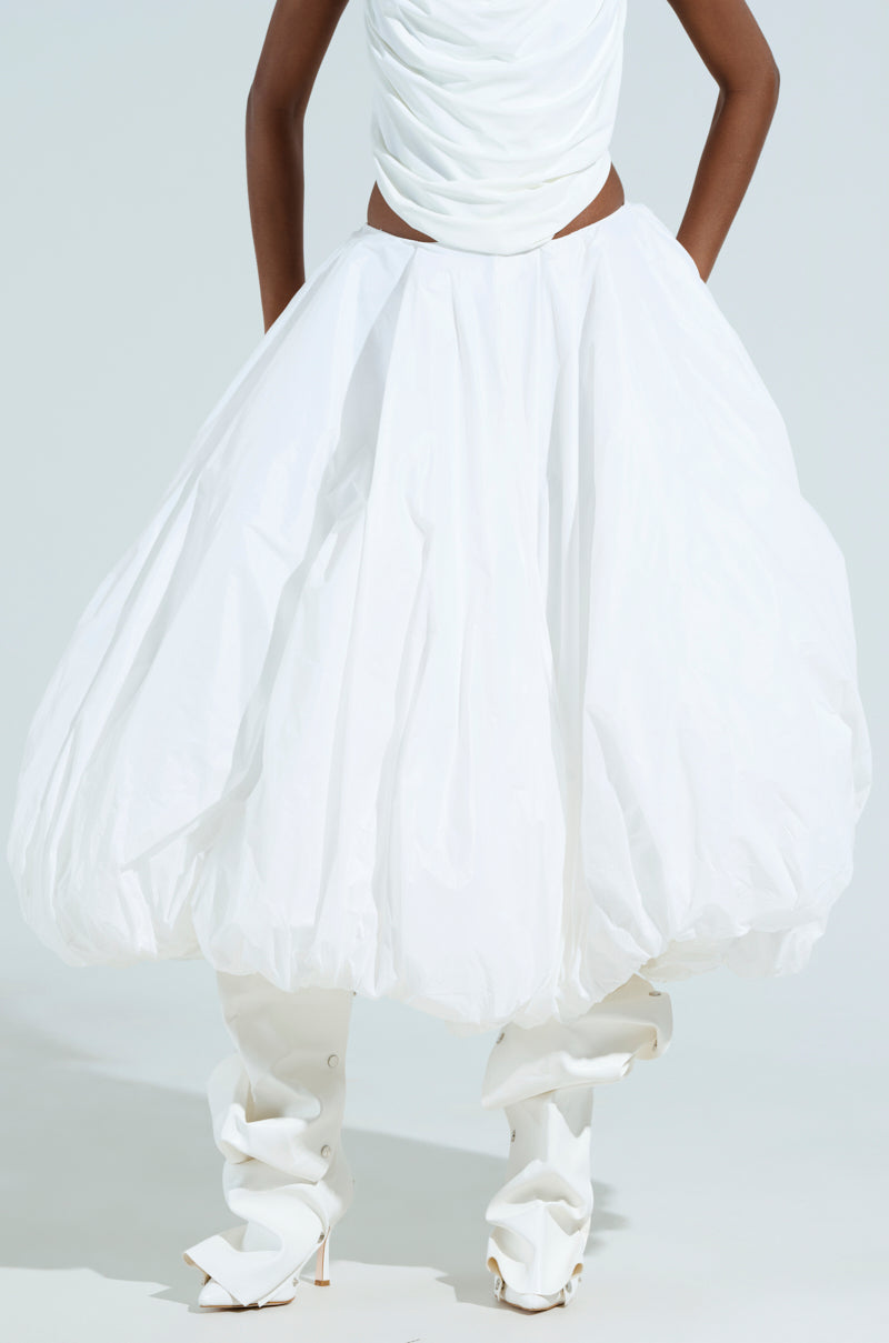 Posing with arms on their hips, the person dons THE BEST MAXI BUBBLE SKIRT paired with a fitted top and white high-heeled boots against a simple, light-colored background.