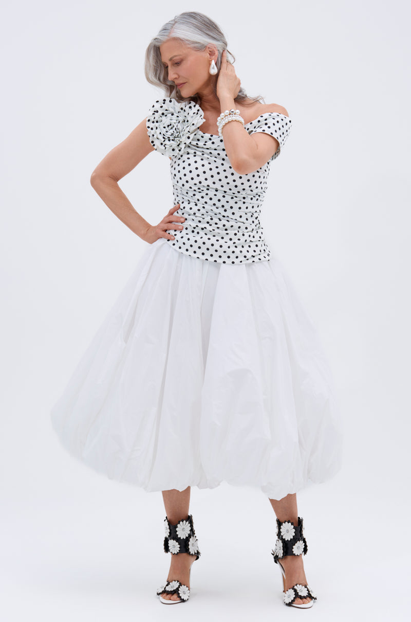 A woman with long gray hair poses gracefully against a white background, wearing THE BEST MAXI BUBBLE SKIRT, a white off-shoulder polka-dot top, statement jewelry, floral heels, and a large floral accessory.