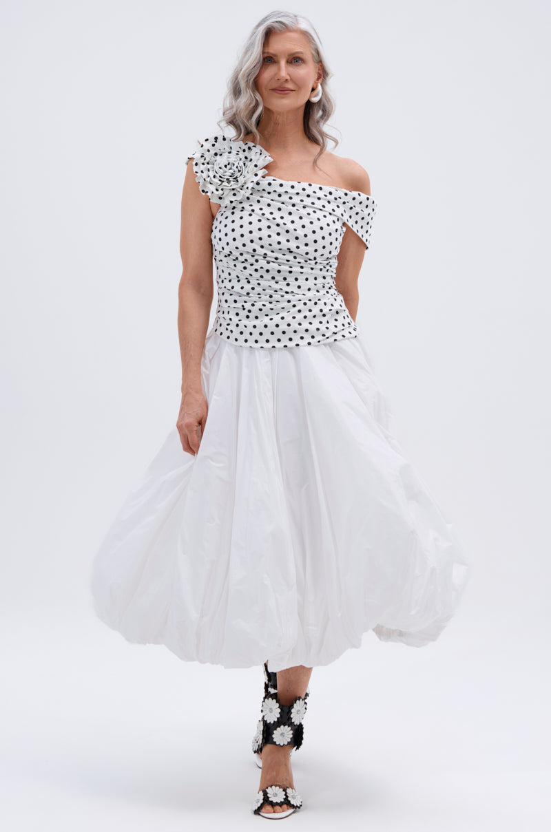 A woman with long gray hair models THE BEST MAXI BUBBLE SKIRT, paired with a white, off-shoulder polka dot top featuring a large floral detail and black-and-white floral sandals, standing against a plain white background.