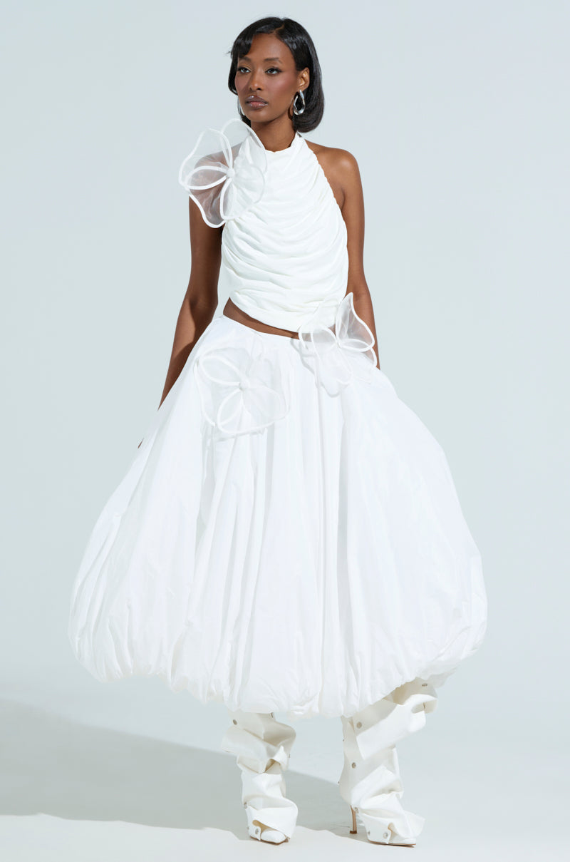 Wearing a chic ensemble, the person features a ruched one-shoulder top paired with THE BEST MAXI BUBBLE SKIRT. The look is adorned with sheer fabric flowers and white knee-high boots, exuding elegance and sophistication against a plain background.