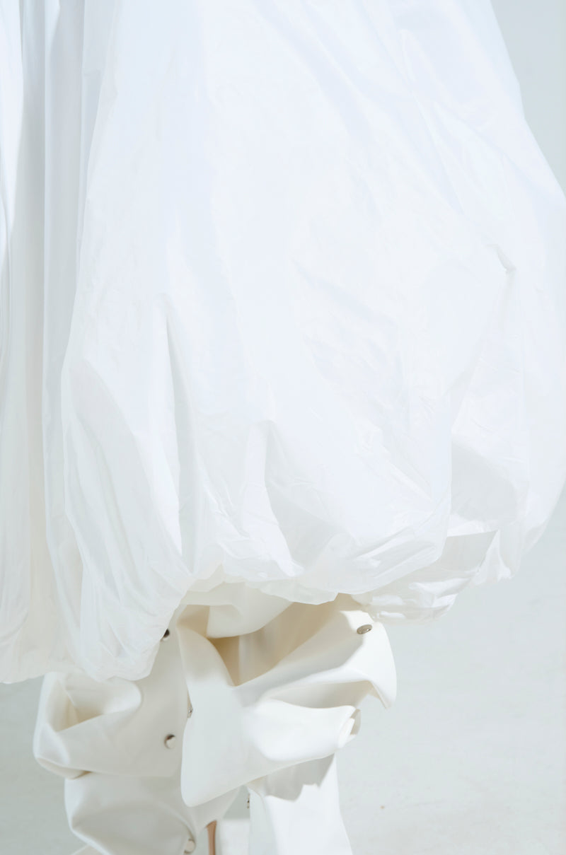 A close-up showcases THE BEST MAXI BUBBLE SKIRT, featuring a billowy, textured white design. Its lightweight, voluminous fabric suggests a midi or bubble skirt style. It covers what may be a leg wrapped in layered white material against a soft, neutral background.