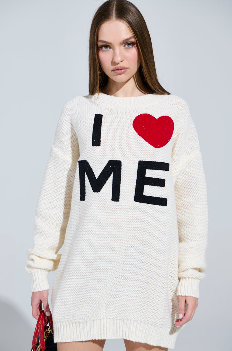 A person with long brown hair effortlessly poses in AKIRA Label's cream "LOVE MYSELF PRINTED SWEATER DRESS," made from stretchy knit fabric, featuring bold black and red "I ❤️ ME" letters, styled with a small red bag against a simple backdrop.