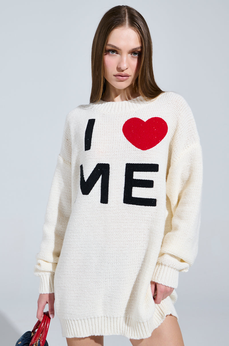 A person with long brown hair dons a LOVE MYSELF PRINTED SWEATER DRESS, displaying "I ♥ ME" in bold black and red. They hold a small red and blue item against a light gray backdrop.