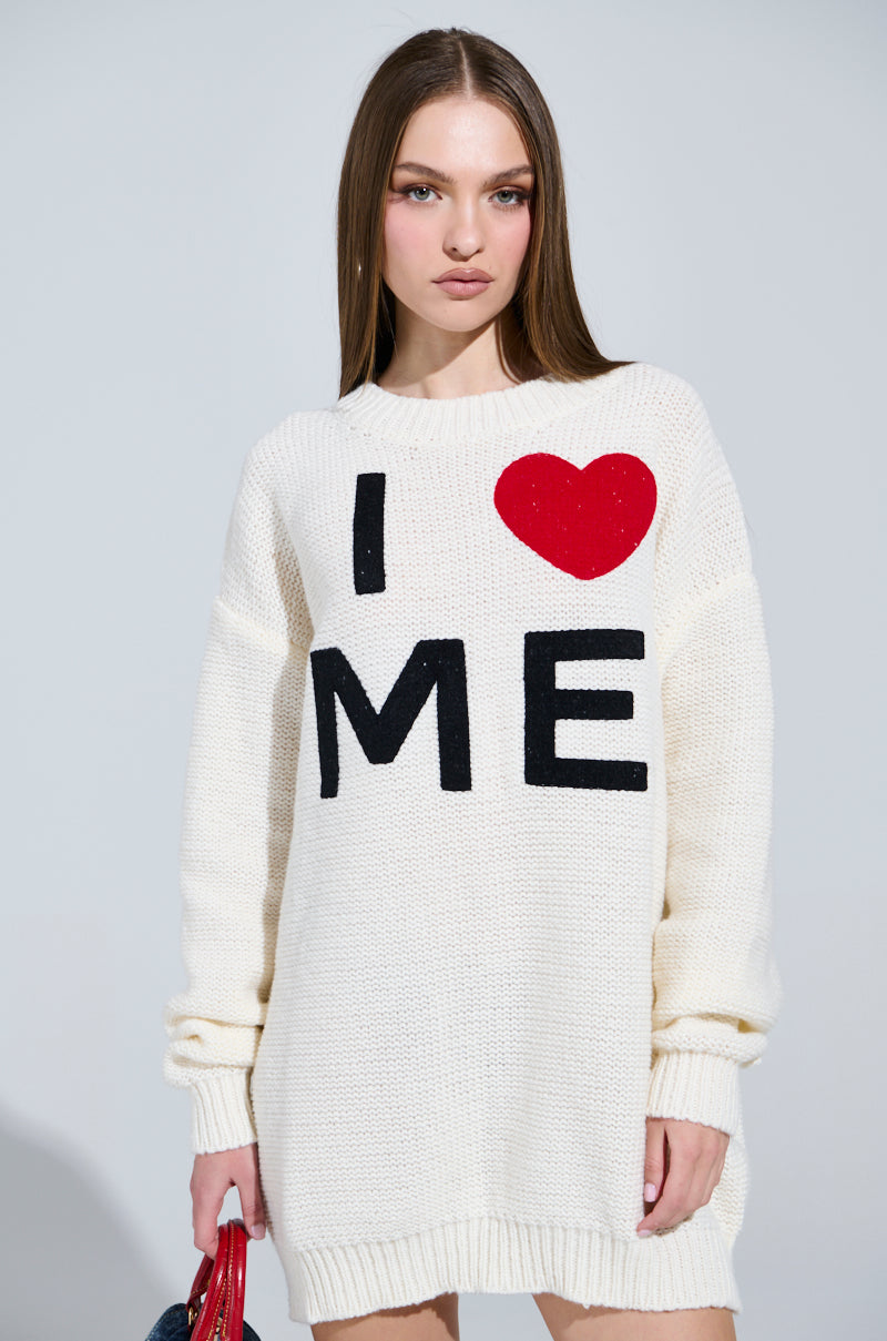 A person with long brown hair wears the LOVE MYSELF PRINTED SWEATER DRESS featuring "I ♥ ME" in bold black and red letters. They hold a red object, stand against a plain background, and gaze directly at the camera with a neutral expression.