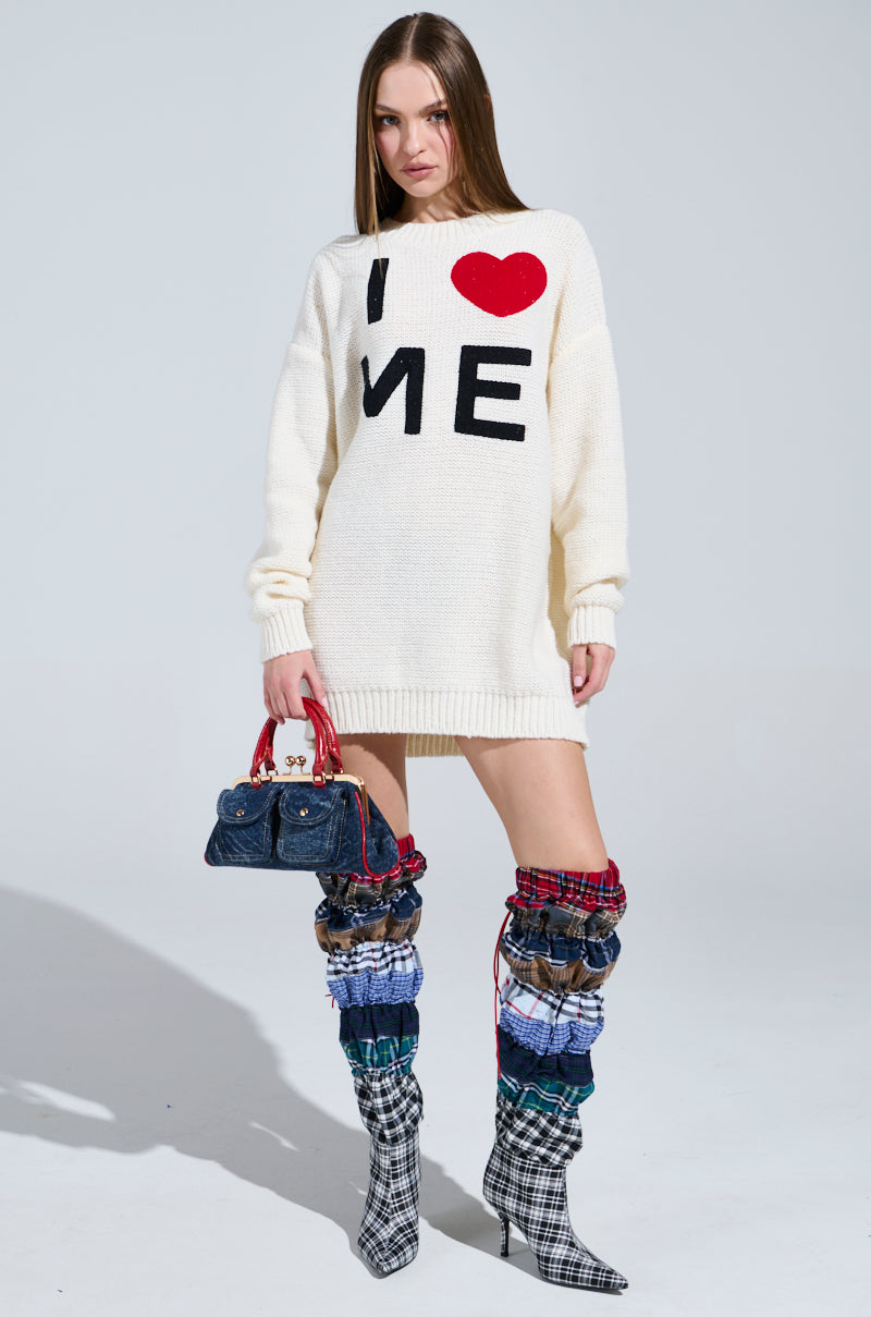 A woman confidently wears a LOVE MYSELF PRINTED SWEATER DRESS made of stretchy knit. She pairs it with a red handbag and AKIRA Label boots, highlighting vibrant fabric layers and black-and-white checkered pointed-toe heels against a simple backdrop.