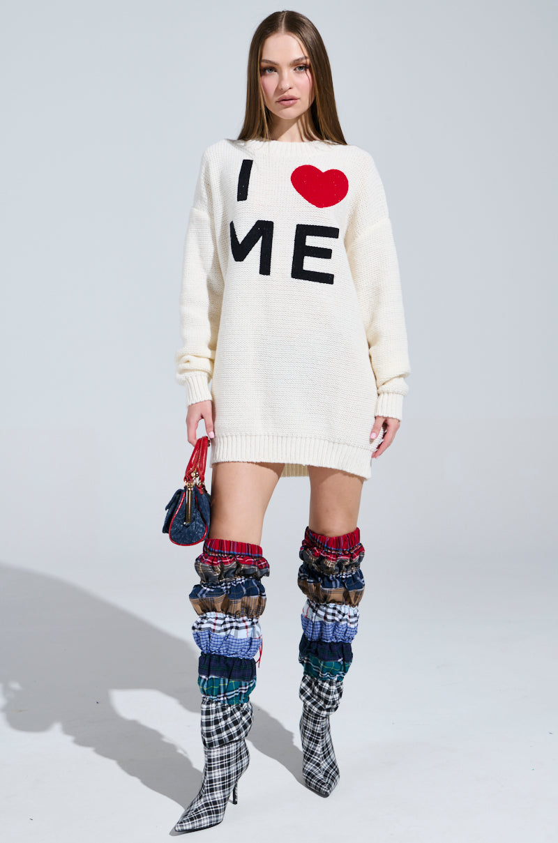 A confident woman wears the cozy LOVE MYSELF PRINTED SWEATER DRESS, paired with colorful mixed-fabric knee-high boots featuring plaid patterns. She accessorizes with a small purse in a well-lit setting that highlights her bold style.