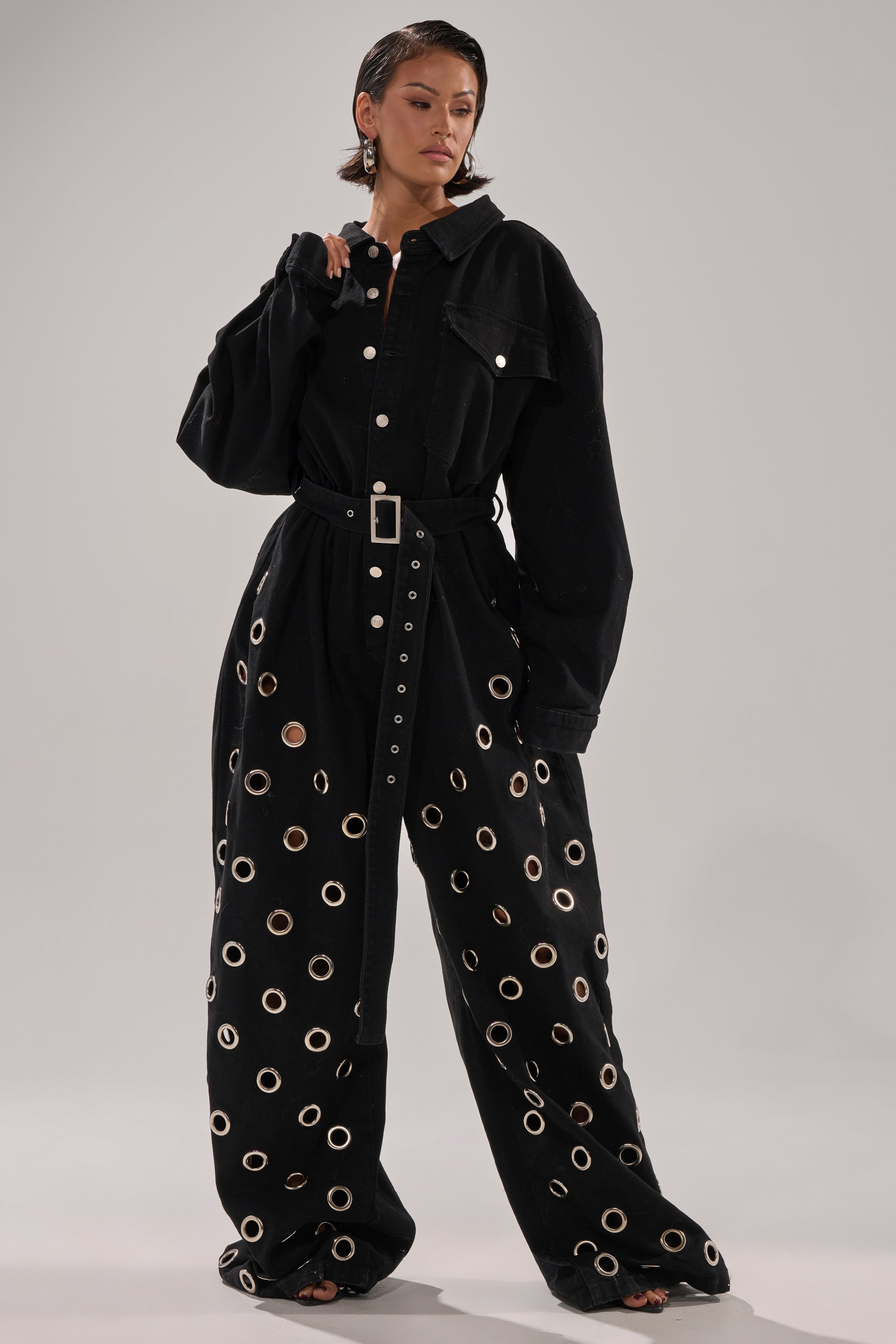 The PLAY YOUR CARDS RIGHT DENIM JUMPSUIT WITH GROMMETS features a black, oversized fit with wide-leg pants, bold silver grommet details, long sleeves, and a matching belt for a modern look. Styled here with hoop earrings and short hair.