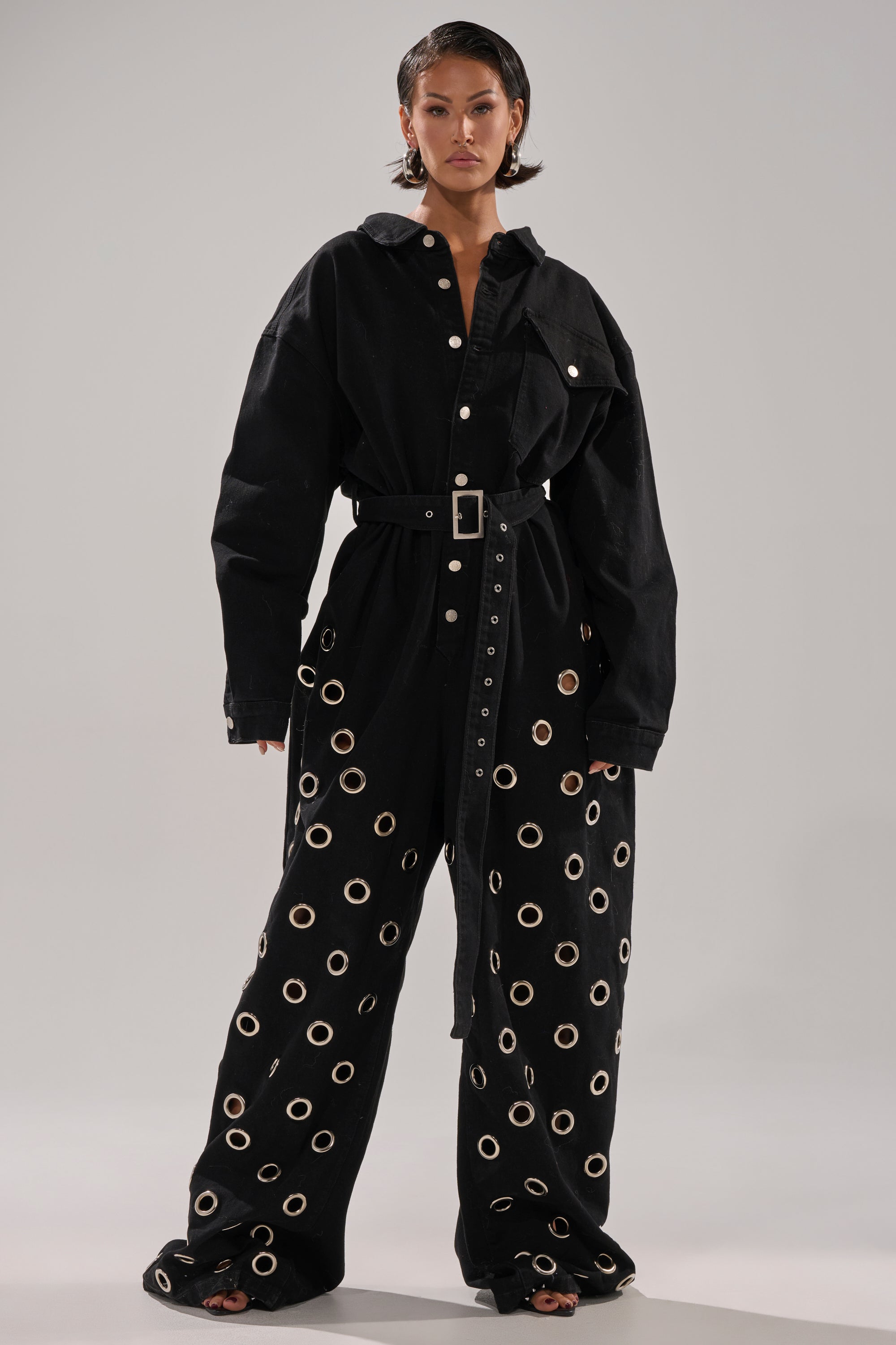 A woman models the PLAY YOUR CARDS RIGHT DENIM JUMPSUIT WITH GROMMETS, featuring a loose fit, belted waist, silver grommet details, and wide legs. She poses confidently against a plain light gray background.