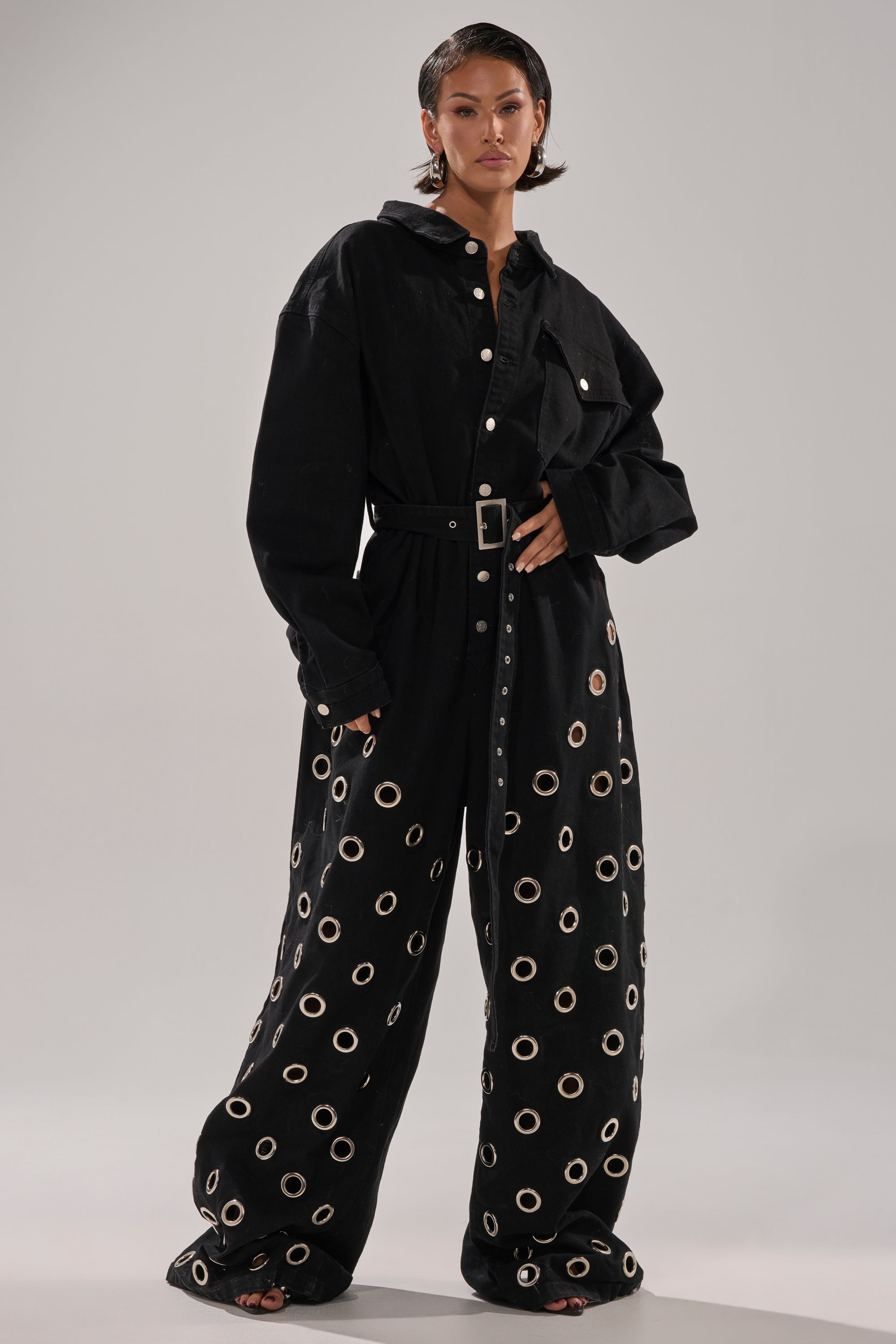 A woman wears the PLAY YOUR CARDS RIGHT DENIM JUMPSUIT WITH GROMMETS—loose-fitting with grommet details, wide-leg pants, belted waist—and black open-toe sandals, standing against a plain background.
