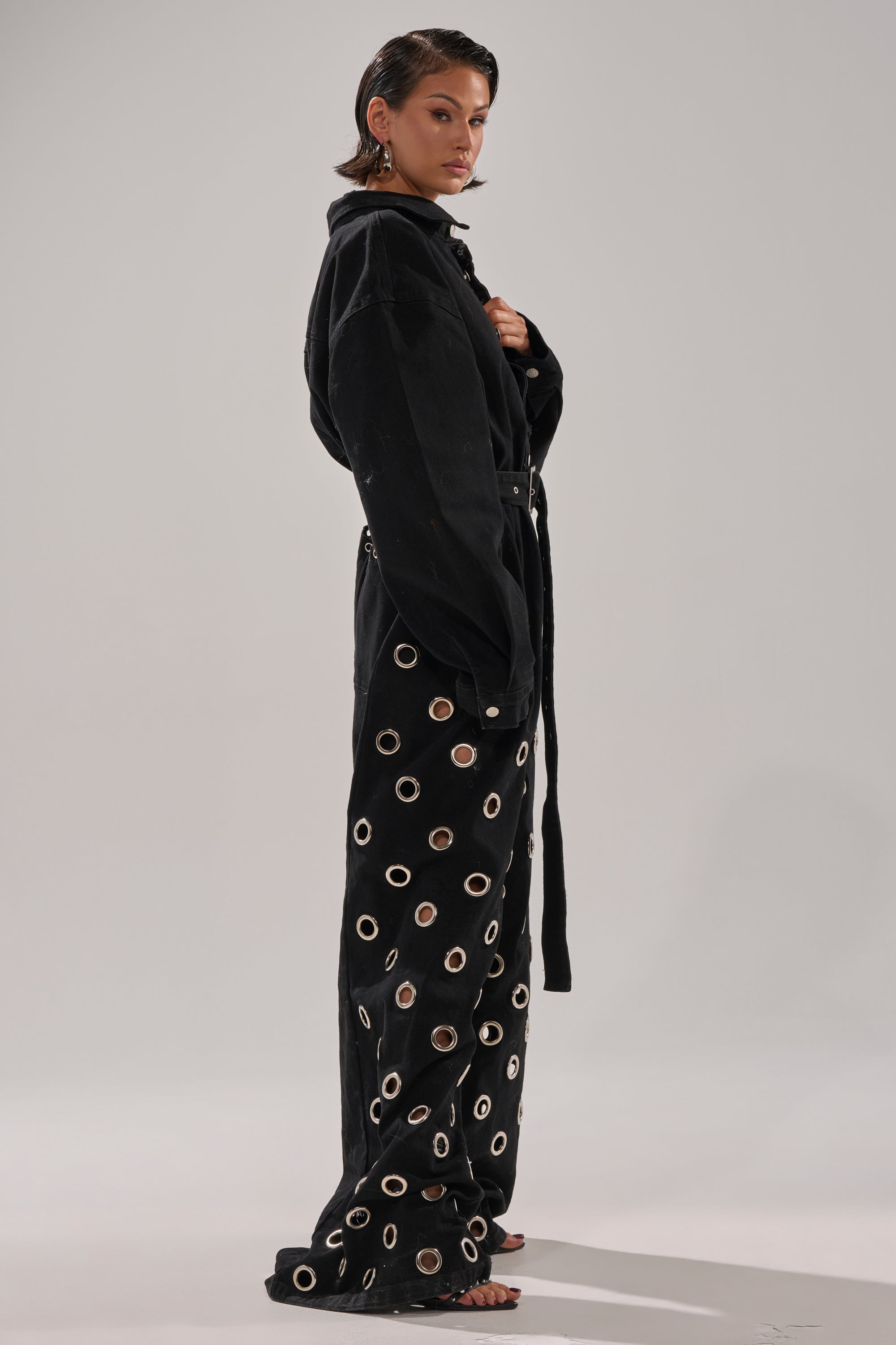 A woman stands barefoot against a plain background, confidently modeling the PLAY YOUR CARDS RIGHT DENIM JUMPSUIT WITH GROMMETS, featuring a black belted design and wide-leg pants with large metallic eyelets. Her wet-look hair is tucked behind her ears.