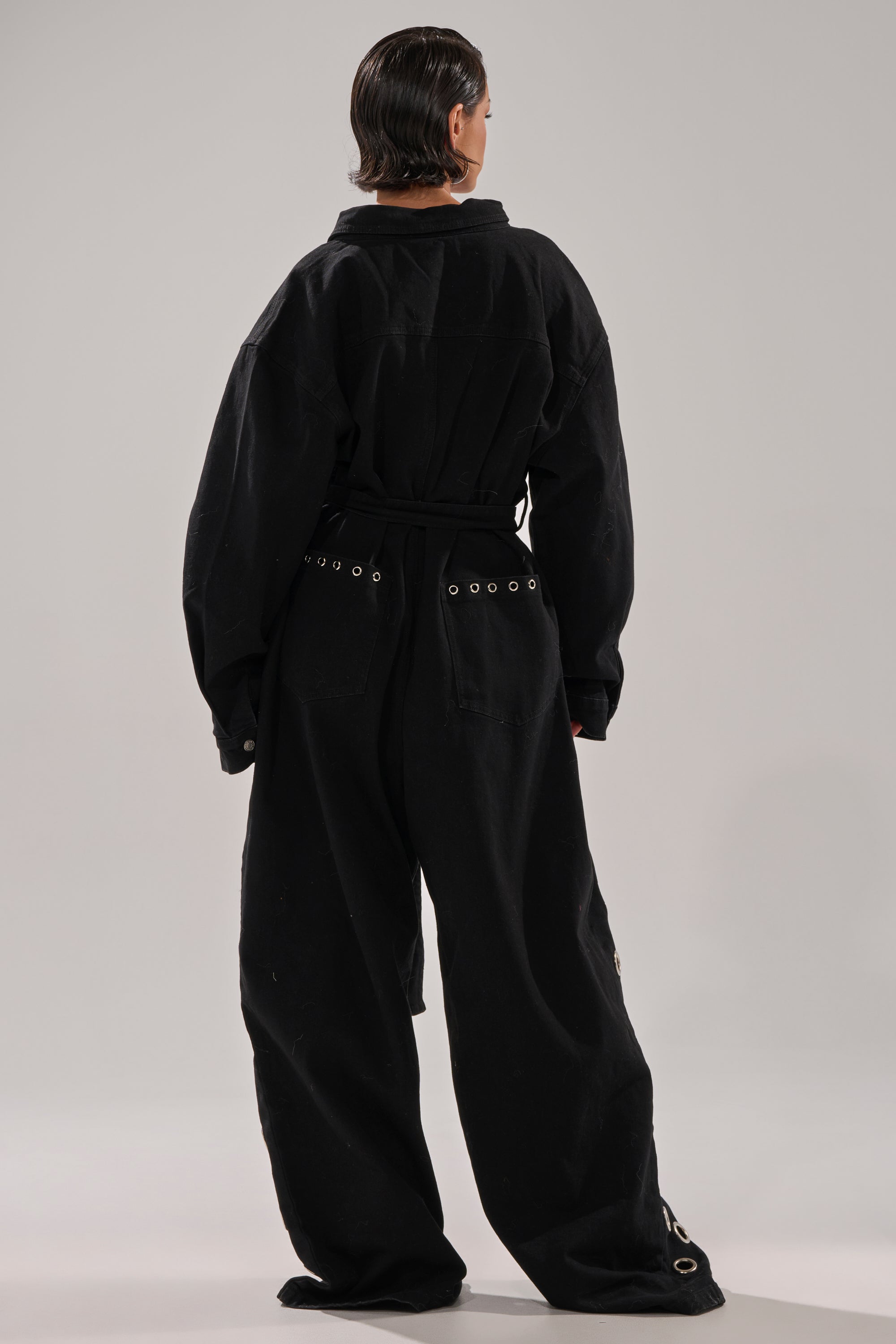 A person faces away, wearing the PLAY YOUR CARDS RIGHT DENIM JUMPSUIT WITH GROMMETS—loose black denim, silver grommet hardware, and a tie belt. Their dark hair is slicked back, set against a plain, light background.