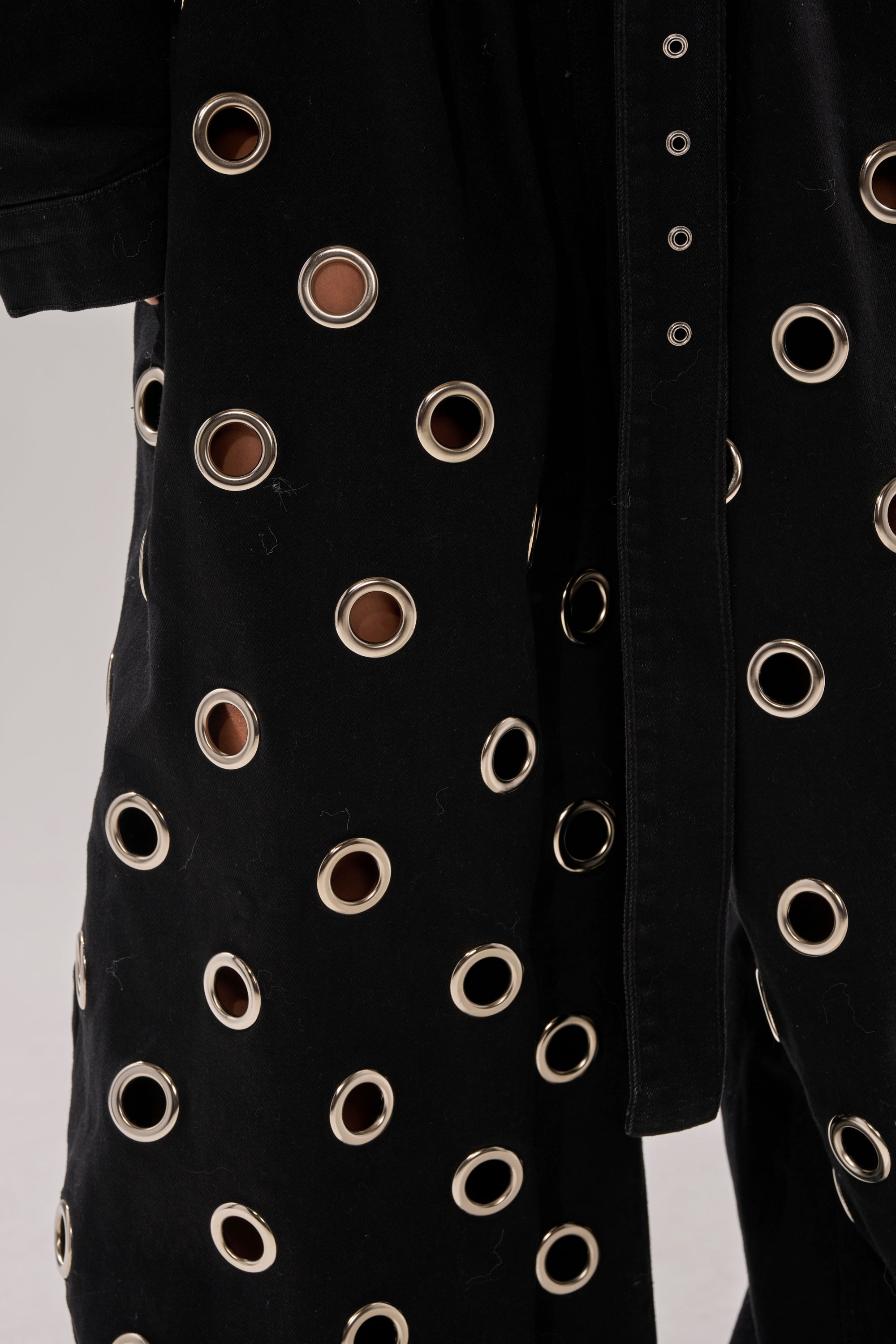 Close-up of the PLAY YOUR CARDS RIGHT DENIM JUMPSUIT WITH GROMMETS shows black fabric with large silver grommets, revealing skin beneath and creating a bold, edgy pattern on this modern twist to classic wide-leg pants.