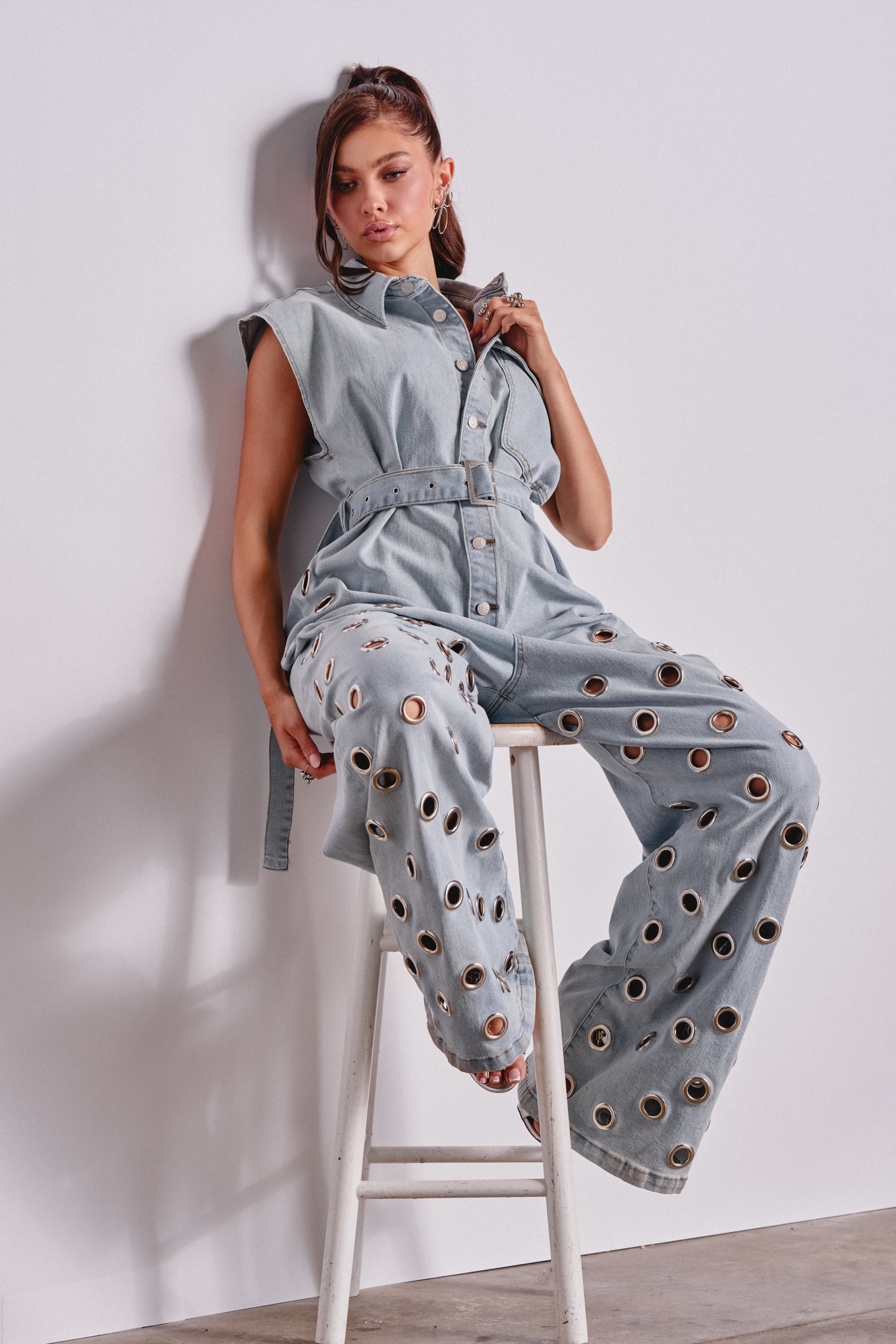 A woman with long hair sits on a white stool against a plain wall, wearing the PLAY YOUR CARDS RIGHT DENIM JUMPSUIT WITH GROMMETS IN LIGHT WASH and hoop earrings. 0