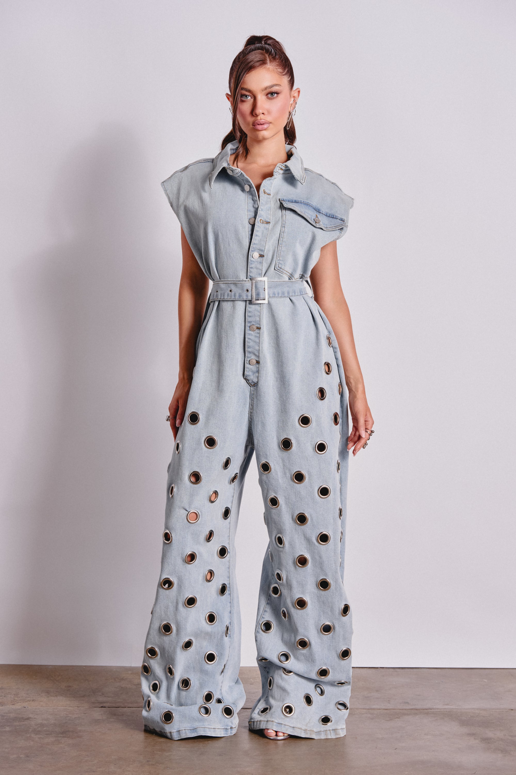 A woman poses against a white background wearing the PLAY YOUR CARDS RIGHT DENIM JUMPSUIT WITH GROMMETS IN LIGHT WASH, featuring cap sleeves, a belted waist, wide legs, and metallic grommet embellishments on the lower half. 2