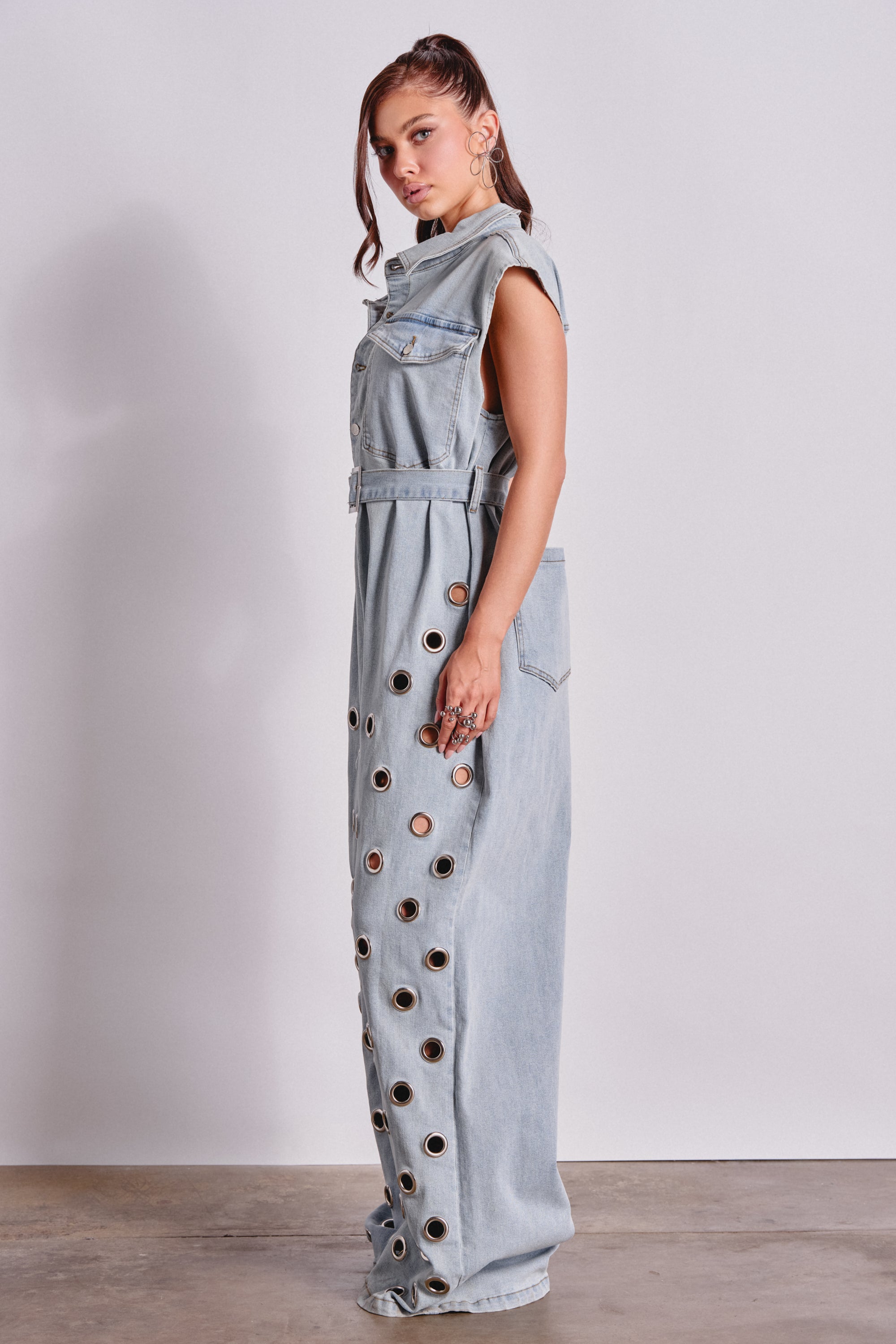 A woman stands sideways in the PLAY YOUR CARDS RIGHT DENIM JUMPSUIT WITH GROMMETS IN LIGHT WASH, featuring a belted waist, front pockets, and metal grommets down the side for an effortlessly chic streetwear look. 3