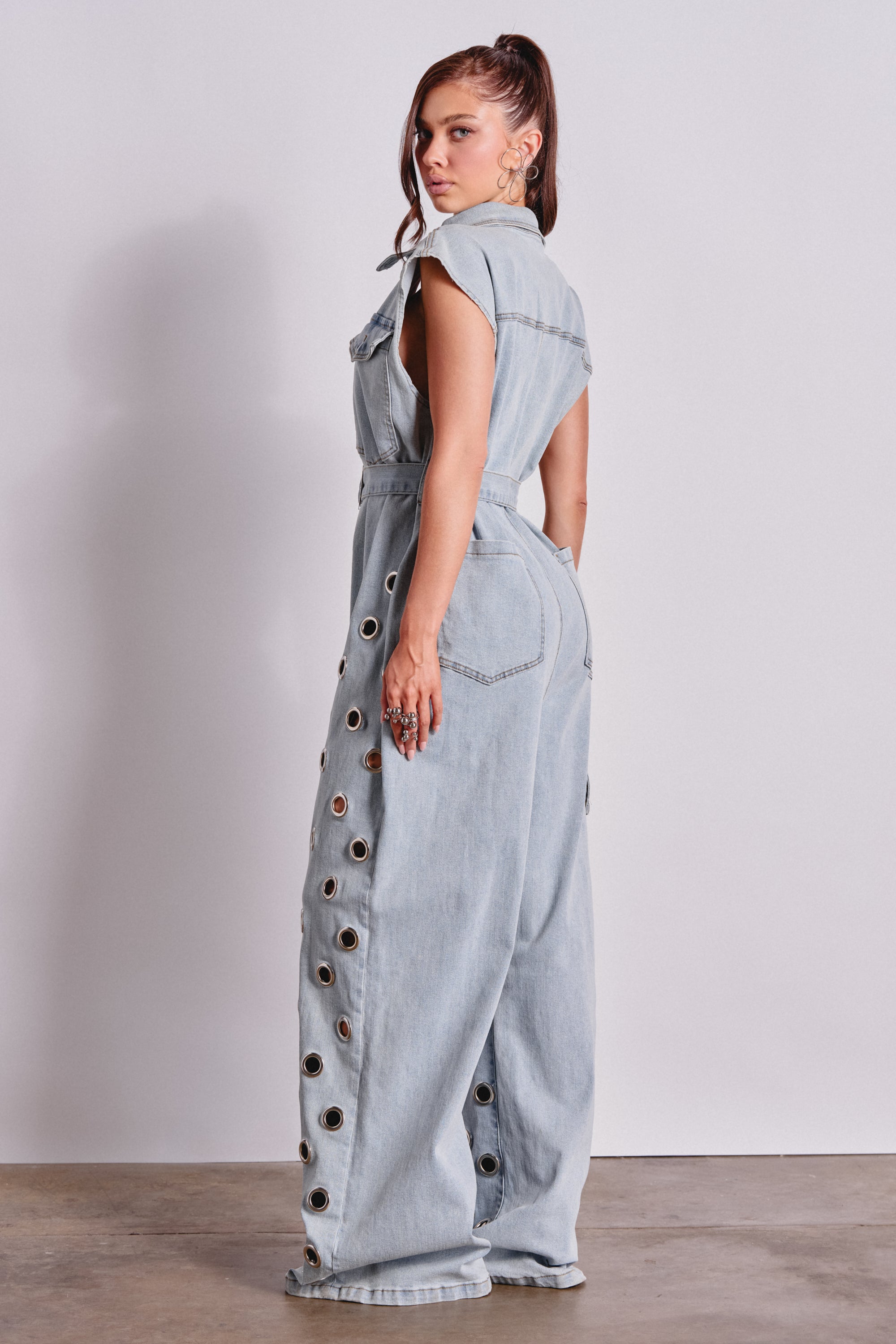 A woman with a high ponytail stands sideways against a light wall, wearing the PLAY YOUR CARDS RIGHT DENIM JUMPSUIT WITH GROMMETS IN LIGHT WASH, featuring wide legs and bold grommet details along the sides. 4