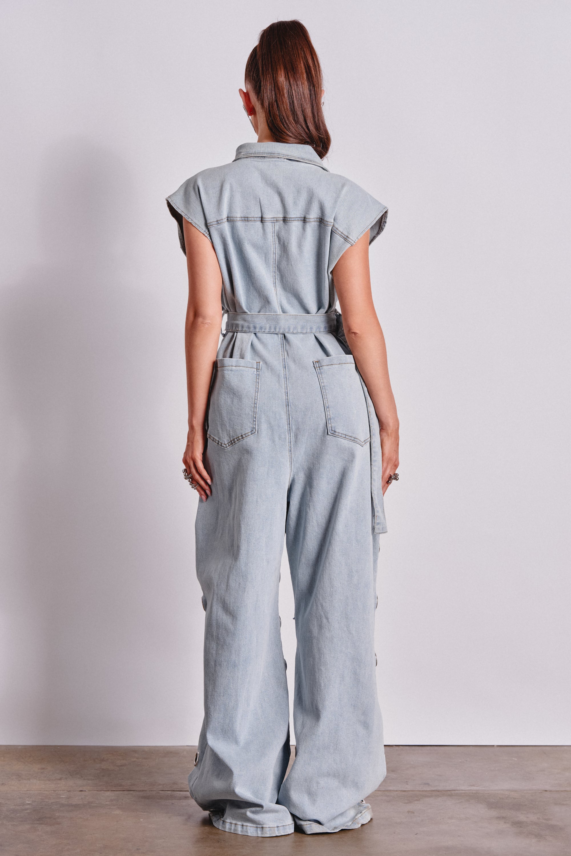 A person with long hair in a ponytail faces away, wearing the PLAY YOUR CARDS RIGHT DENIM JUMPSUIT WITH GROMMETS IN LIGHT WASH—featuring wide legs and chic seam details—styled as modern streetwear against a plain white background. 5