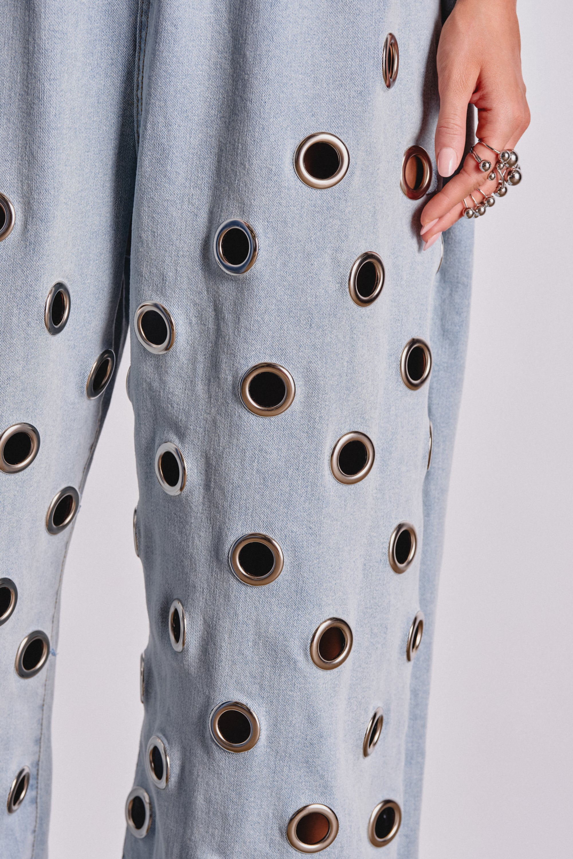 A close-up of a person in the PLAY YOUR CARDS RIGHT DENIM JUMPSUIT WITH GROMMETS IN LIGHT WASH, featuring bold metallic grommets. Their ringed hand and beaded bracelet enhance this striking and modern streetwear romper look against a plain background. 6