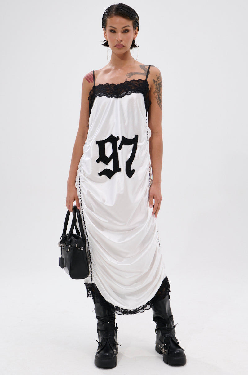 A person stands facing forward in the GO SPORTS RUCHED MIDI DRESS, a white satin varsity-style piece with black lace trim and the number "97" on the front. They have tattoos, wet-look hair, black boots, and hold a black handbag against a plain white background.