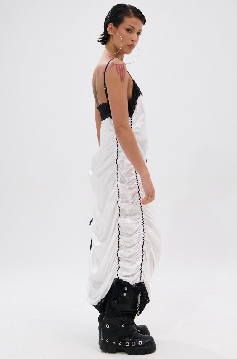 A person with short dark hair and large hoop earrings models the GO SPORTS RUCHED MIDI DRESS in white with black lace trim, styled with chunky black platform boots, standing sideways against a plain white background.