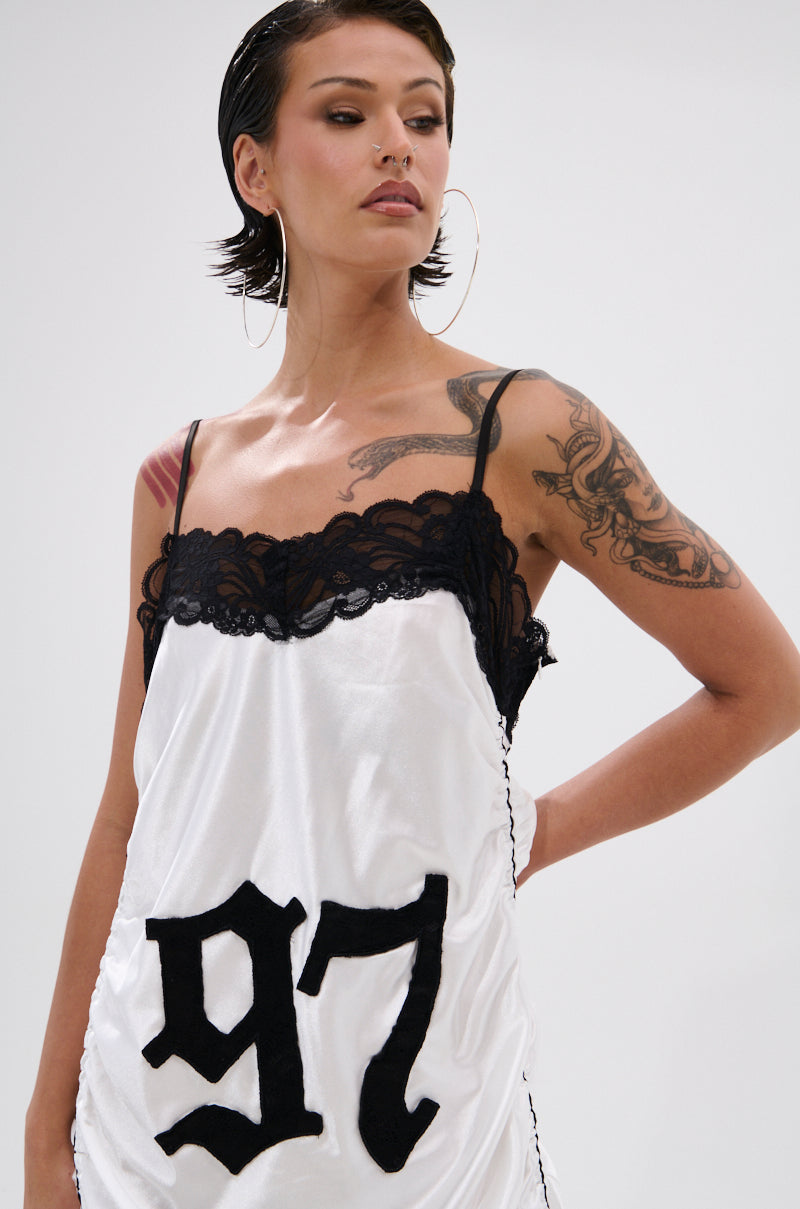A woman with short dark hair and hoop earrings models the GO SPORTS RUCHED MIDI DRESS, a white varsity-style satin slip dress with black lace trim and bold "97" on the front. She displays tattoos on her right shoulder and arm.