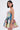 A woman with straight dark hair wears the STAND OUT BROCADE MINI DRESS—a colorful, floral mini dress with a flared skirt and thin straps—as she stands sideways, looking at the camera against a plain light background, accessorized with large hoop earrings.