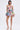 A woman with straight dark hair stands facing forward in the STAND OUT BROCADE MINI DRESS, featuring a sleeveless metallic design with thin straps and a flared skirt. She pairs it with gold high-heeled sandals against a plain light background.