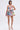 A woman wears the STAND OUT BROCADE MINI DRESS, featuring a fitted bodice and flared skirt in colorful patchwork. She pairs it with gold strappy high-heeled sandals and has straight, shoulder-length hair against a plain white background.