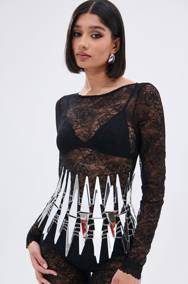 A woman with a sleek bob wears a sheer black lace top and high-waisted bottoms, accessorized with the DONT GET CLOSE MIRROR BELT and silver earrings, standing against a plain light background.