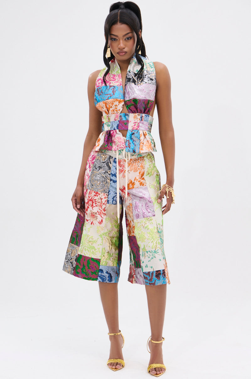 A person wearing a vibrant, sleeveless patchwork jumpsuit with a high-rise waistband and colorful patterns, paired with gold sandals and a matching bracelet, exudes effortless style against a plain white backdrop. The look is highlighted by the MIXED FEELINGS BROCADE BERMUDA SHORT.