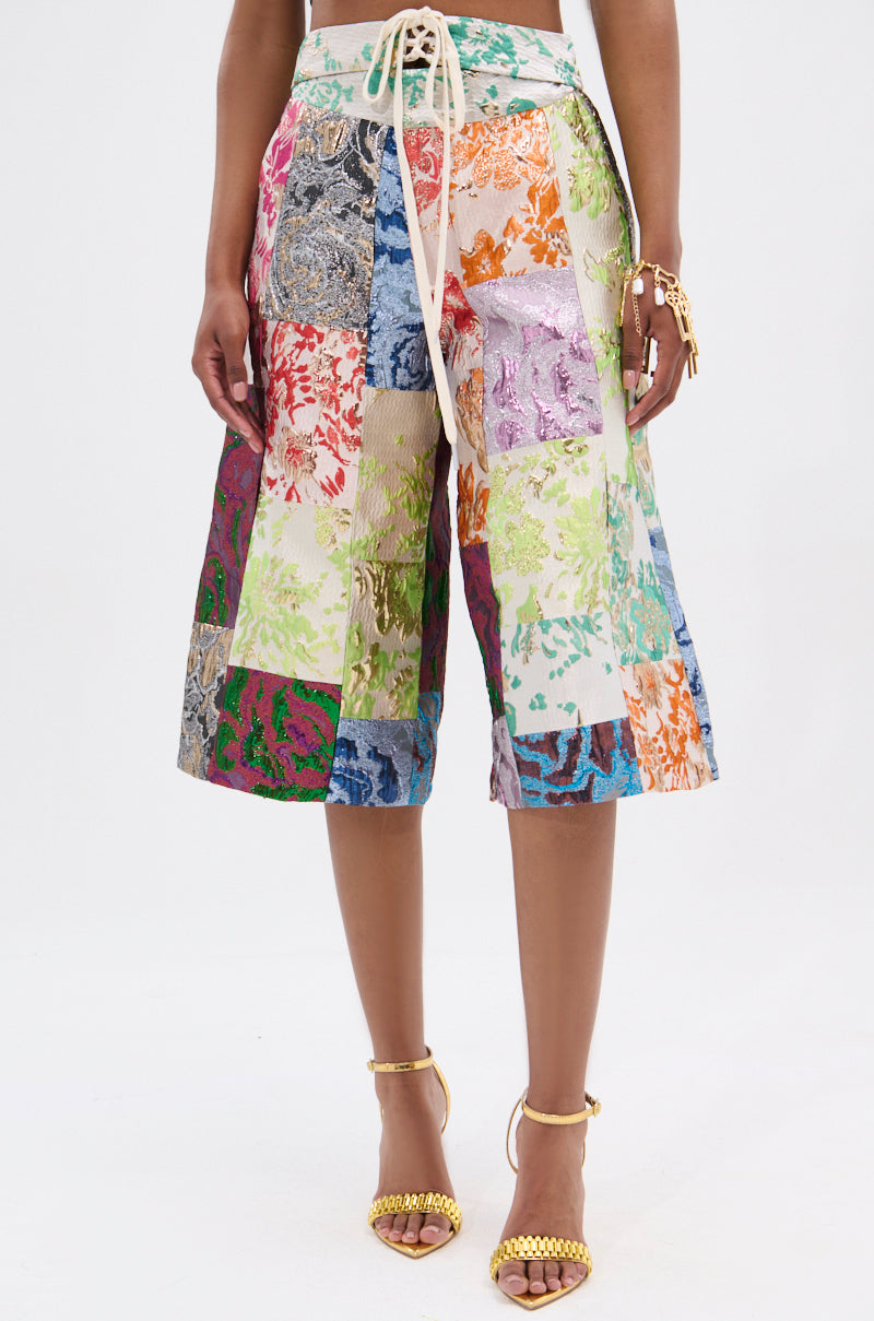 The person wears the MIXED FEELINGS BROCADE BERMUDA SHORT featuring a patchwork of colorful fabric squares with a high-rise drawstring waistband, paired with strappy gold high heels and a gold bracelet against a plain white background.