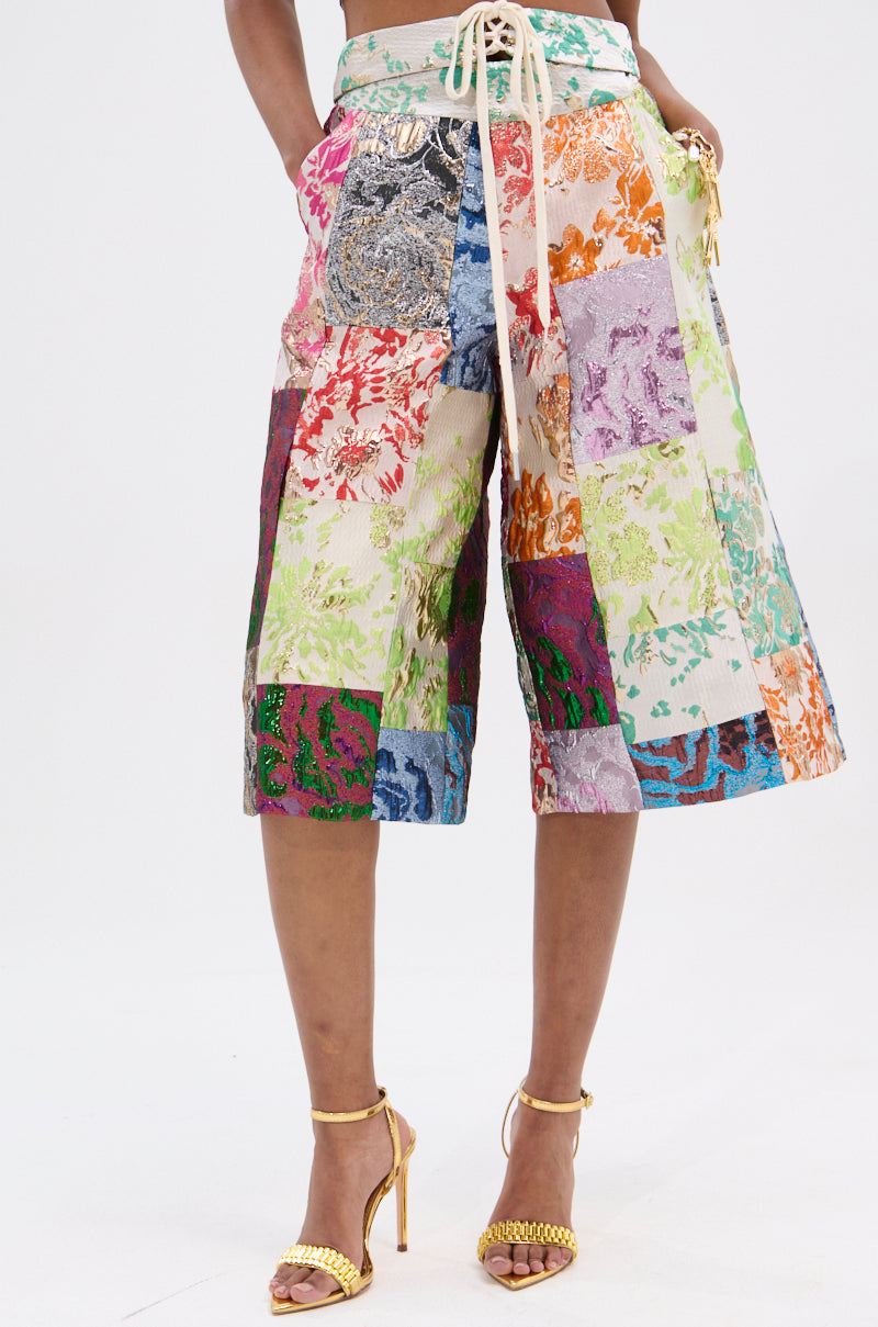 Someone wears the vibrant "MIXED FEELINGS BROCADE BERMUDA SHORT," featuring floral patchwork, a high-rise drawstring waist, and fringe detail on a neutral base. Paired with gold high-heeled sandals, these knee-length shorts make a bold statement.
