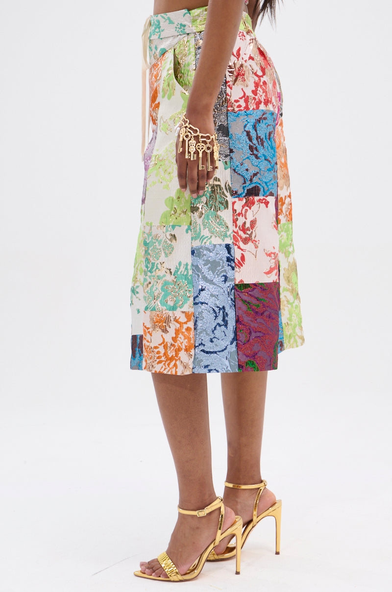 Side view of a person displaying a colorful MIXED FEELINGS BROCADE BERMUDA SHORT with a high-rise waistband, paired with gold high heels and a decorative gold hand accessory featuring key-shaped elements against a plain white background.