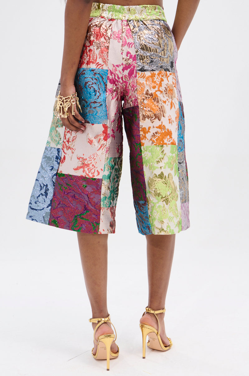Wearing the vibrant MIXED FEELINGS BROCADE BERMUDA SHORT with a floral patchwork in pink, blue, and green hues, paired with strappy gold heels and a chunky gold bracelet creates an eye-catching look against the plain white background.