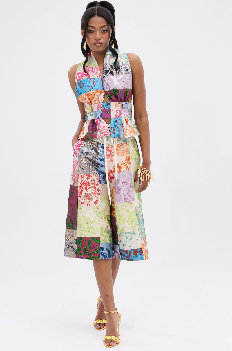 A person in a sleeveless patchwork dress with vibrant patterns stands against a plain background. Their long hair is styled in ponytails, paired with yellow heels and gold accessories, accompanied by high-rise waistband MIXED FEELINGS BROCADE BERMUDA SHORTS.