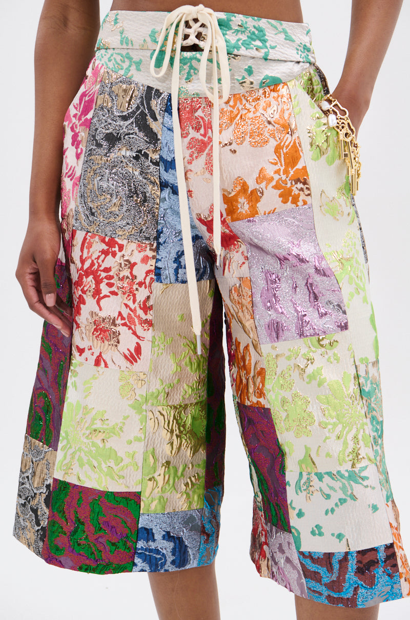 A person flaunts the MIXED FEELINGS BROCADE BERMUDA SHORT with vibrant, floral patchwork and sequins. This high-rise short boasts a lace-up front, metallic fabric in red, green, blue, purple, and gold hues, with a side keychain accessory.