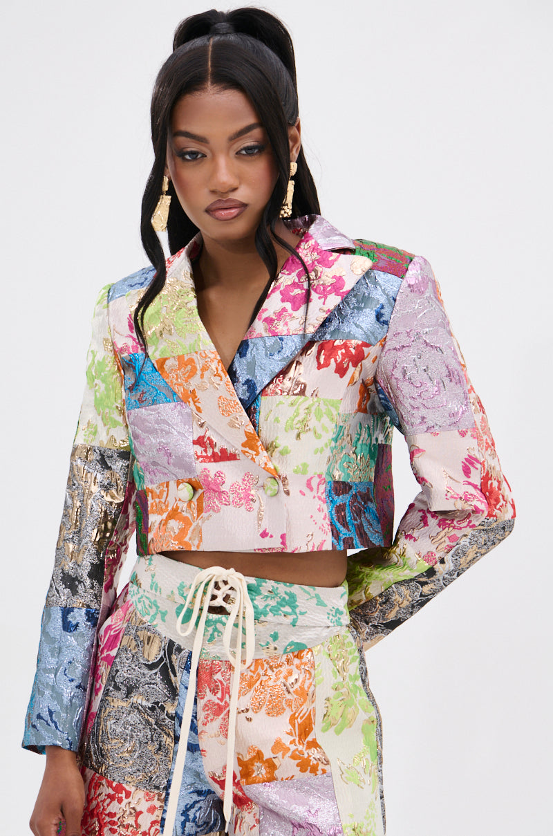 A woman in a MIXED FEELINGS BROCADE BLAZER and matching floral patchwork pants stands against a plain background, showcasing vibrant multicolored designs. Her long dark hair and gold earrings enhance her chic look.